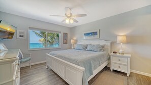 Ein Schlafzimmer mit Bett, Schreibtisch, Stuhl, Nachttisch, Lampe, Deckenventilator und einem Fenster mit Meerblick.