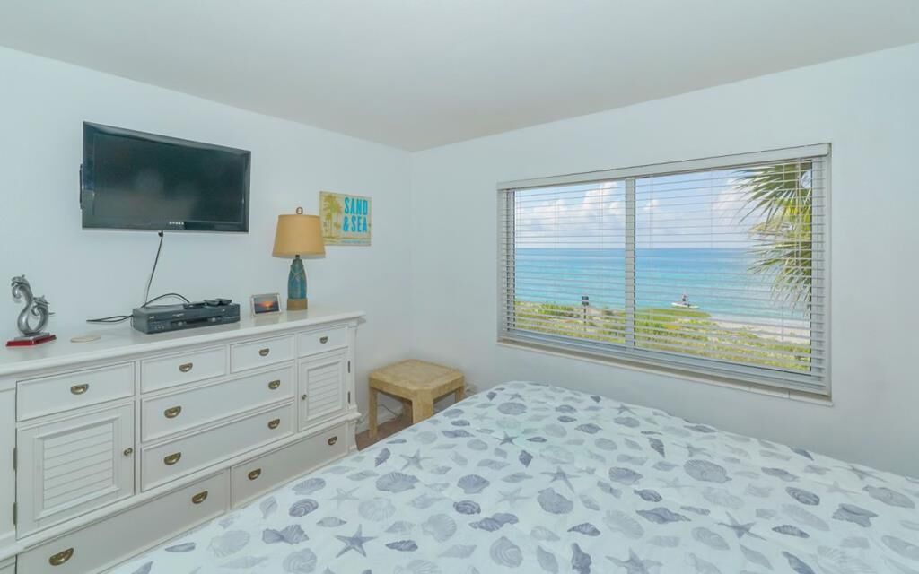 Une chambre à coucher comprenant un lit, une télévision, une commode, une lampe, un petit tabouret et une fenêtre donnant sur la plage.
