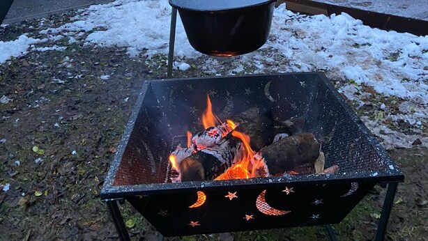 Un foc de llar amb llenya encès, una olla penjant i una taula amb cadires davant d'un edifici.