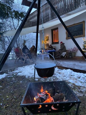 Un foyer avec des bûches qui brûlent, un pot suspendu et une table avec des chaises devant un bâtiment.