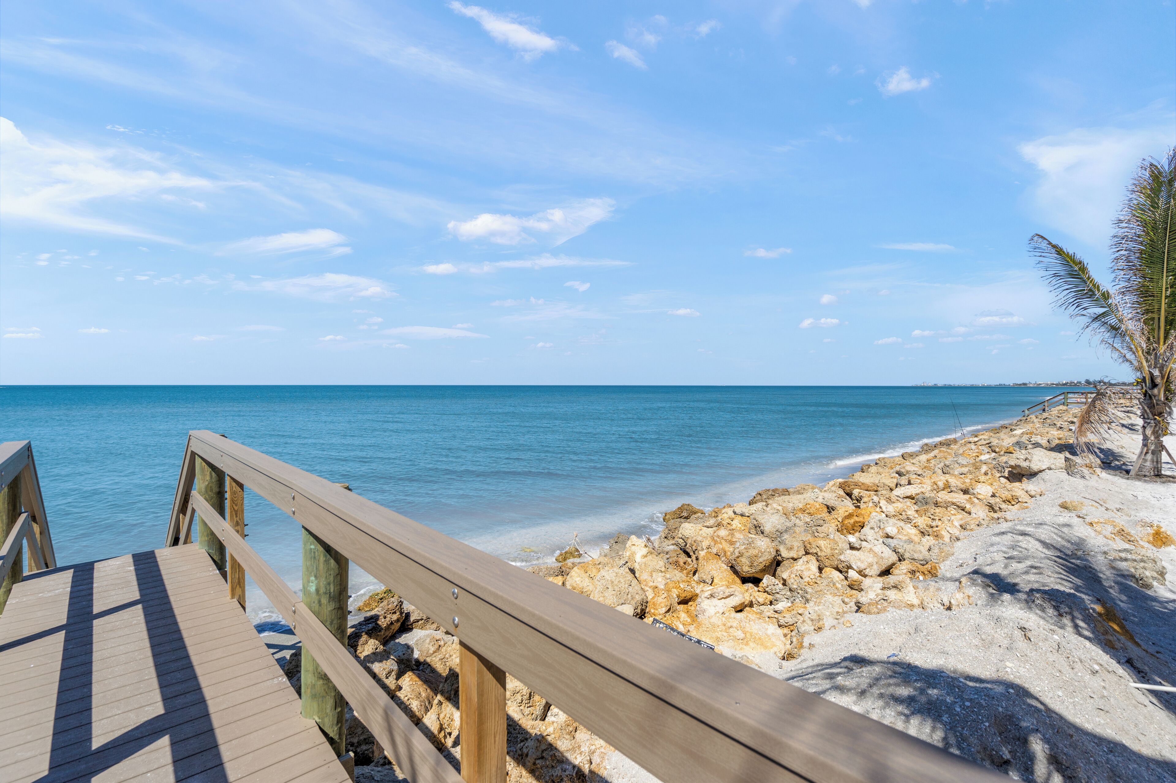 Una passerella in legno che conduce a una spiaggia rocciosa con acqua cristallina e una palma.