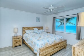 Une chambre avec un lit en bois, un ventilateur de plafond, une table de chevet avec une lampe et une fenêtre donnant sur l’océan.
