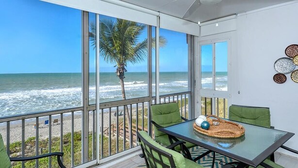A balcony with a glass door overlooking the beach, a table with chairs, and decorative wall art. - Your Slice of Gulf-Front Paradise (Sarasota)
