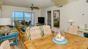 A room with a wooden dining table, wicker chairs, a television, and a view of the ocean through large windows. - Your Slice of Gulf-Front Paradise (Sarasota)