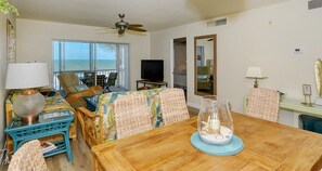 A room with a wooden dining table, wicker chairs, a television, and a view of the ocean through large windows. - Your Slice of Gulf-Front Paradise (Sarasota)
