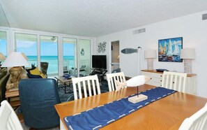 A room with a wooden dining table, white chairs, a TV, and a view of the ocean through large windows. - Unobstructed Panoramic Gulf Views & Steps to the Shoreline (Sarasota)