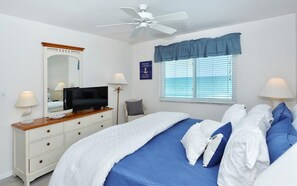 Een slaapkamer met een bed, een televisie, een plafondventilator, een raam met zonwering en een spiegel.