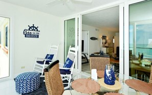 A room with a glass door leading to a balcony, white chairs, a patterned ottoman, and a wall sign. - Unobstructed Panoramic Gulf Views & Steps to the Shoreline (Sarasota)