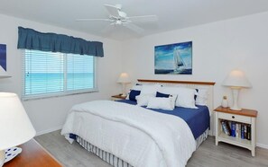 A bedroom with a bed, a ceiling fan, a window with blinds, a nightstand with a lamp, and a painting on the wall. - Unobstructed Panoramic Gulf Views & Steps to the Shoreline (Sarasota)