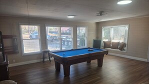 Een pooltafel in een kamer met grote ramen, houten vloeren en een plafondventilator.