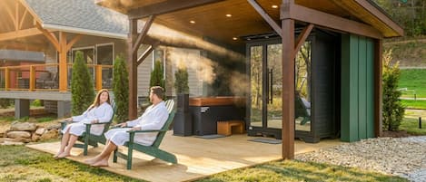 A covered outdoor seating area with a grill, two people sitting on chairs, and a view of a house in the background.