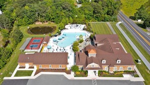 Vista aérea de um bairro residencial com uma piscina grande cercada por casas, uma quadra de tênis e uma estrada.