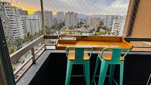Ein Balkon mit einem Holztisch und zwei grünen Stühlen, mit Blick auf eine Stadtlandschaft mit Hochhäusern.