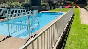 Ein rechteckiger Swimmingpool mit einem Metallzaun, umgeben von einem gepflegten Rasen und einer Spielanlage für Kinder im Hintergrund.