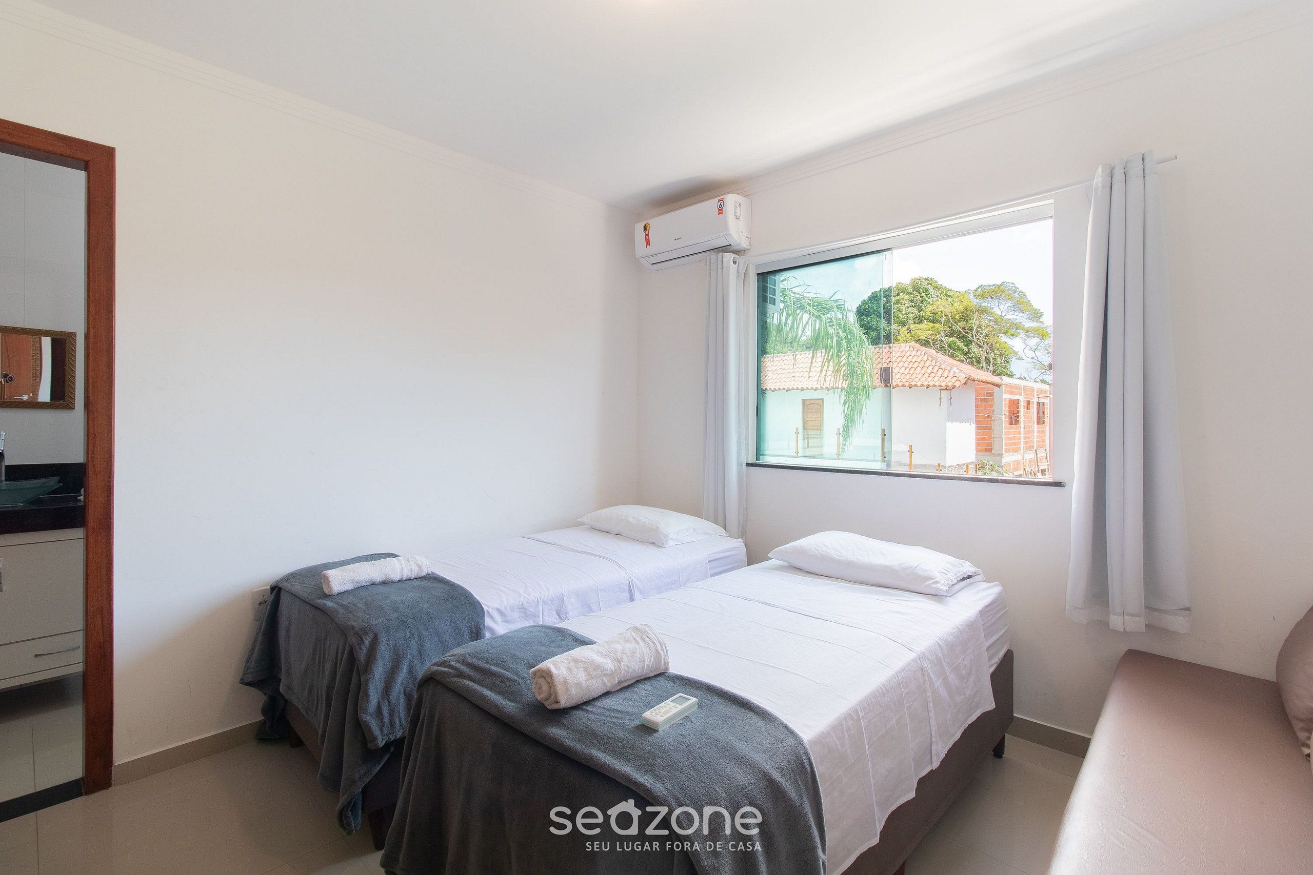 A room with two beds, a window with a view of buildings, and an air conditioning unit.