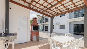 An outdoor patio with a brick oven, white plastic chairs, and a table. - Apts in Condominium w/ Pool and Barbecue - Ctys (Porto Seguro)