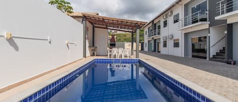 A swimming pool with a pergola, a tiled deck, and a building with balconies.