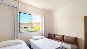 A hotel room with two single beds, a sofa, a large window with a view of a building, and a wall-mounted air conditioner. - Apts in Condominium w/ Pool and Barbecue - Ctys (Porto Seguro)