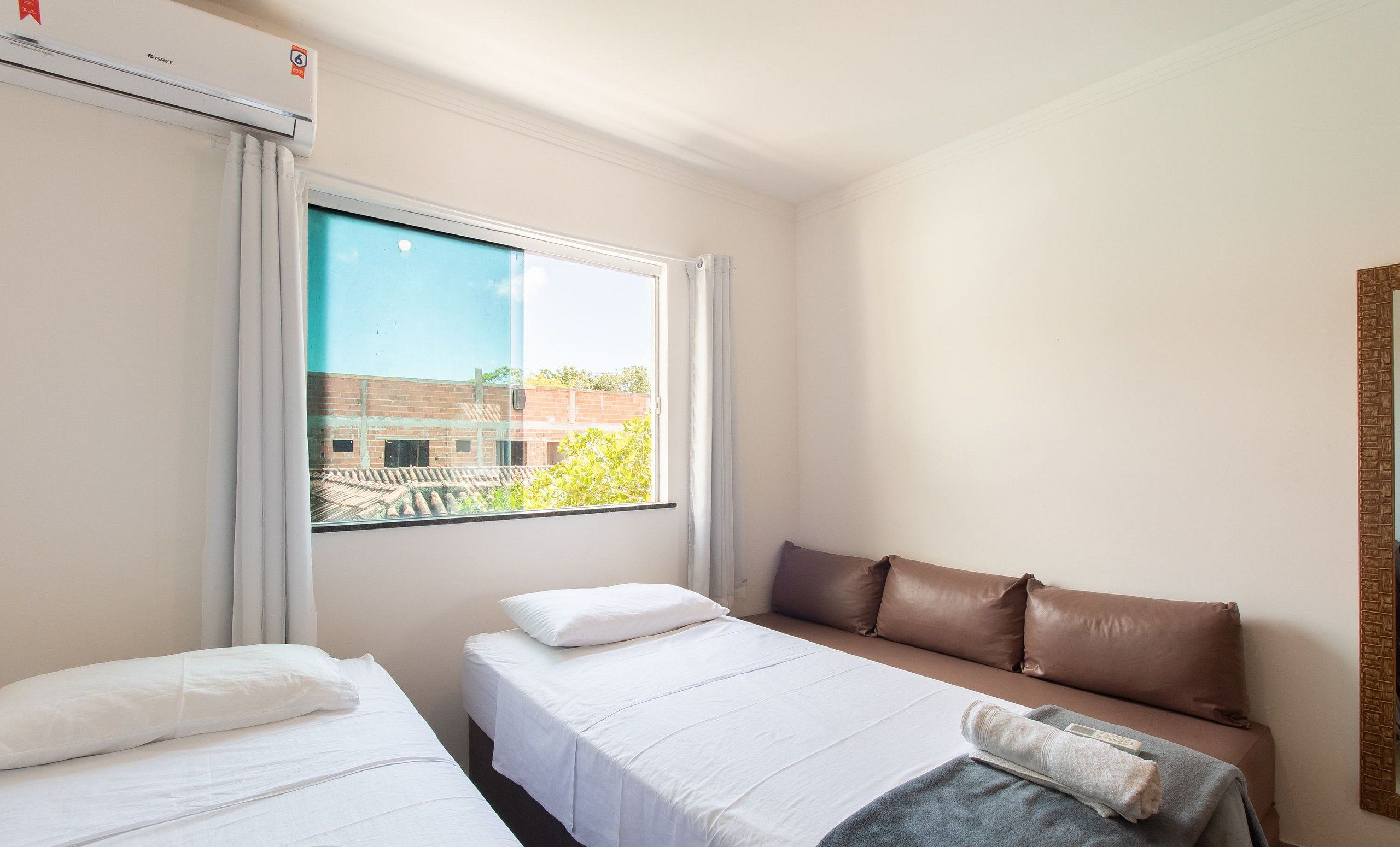 A hotel room with two single beds, a sofa, a large window with a view of a building, and a wall-mounted air conditioner.
