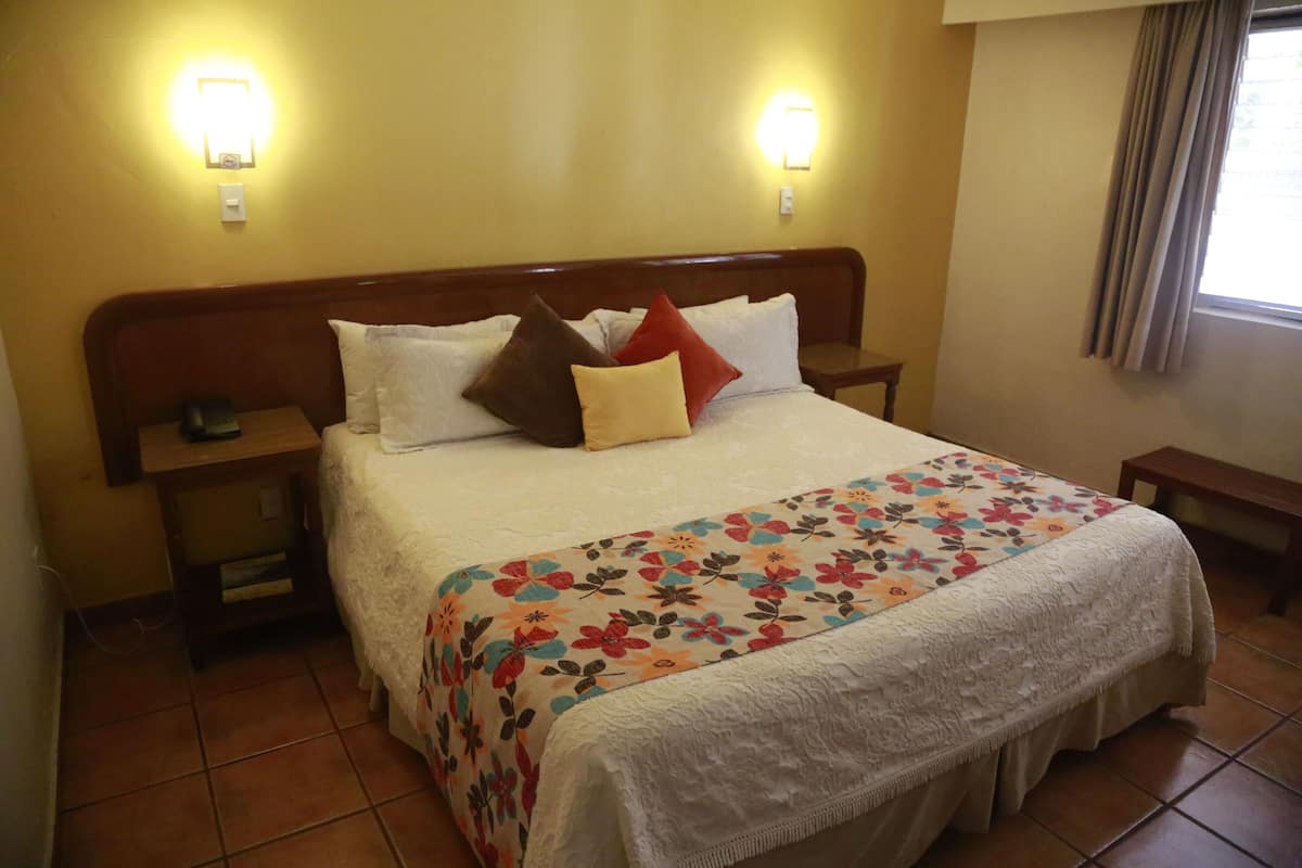 A hotel room with a large bed, two bedside tables, a nightstand, a floral bedspread, and two wall-mounted light fixtures.