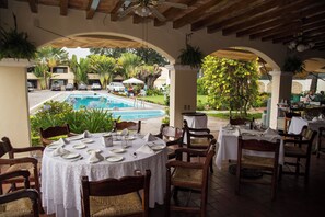 Un área de comedor al aire libre con mesas redondas, sillas y una piscina al fondo.