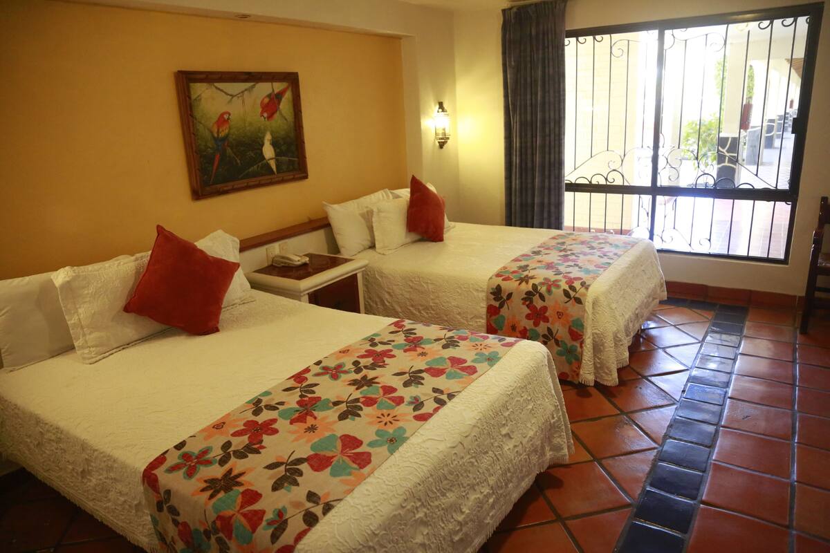 A hotel room with two beds, each with a floral bedspread and red pillows. There is a painting of parrots on the wall, a nightstand with a phone, and a window with a view of the outside.