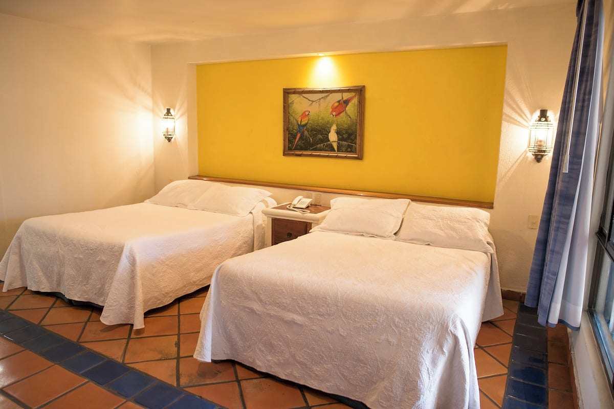 A hotel room with two single beds, a nightstand with a phone, and a painting of birds on the wall.