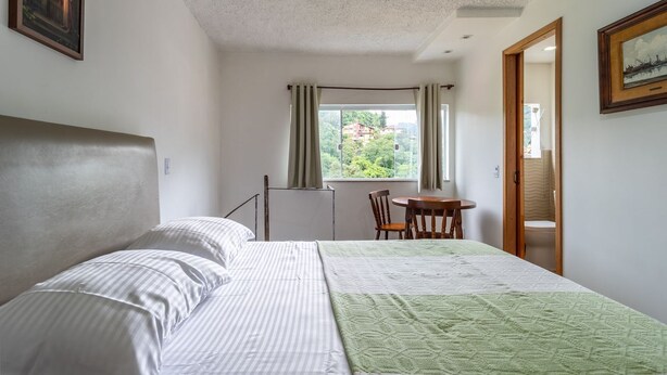 A bedroom with a bed, a chair, a small table, and a window with curtains. - Apto Para 3 Pessoas na Rua Teresa (Petrópolis)