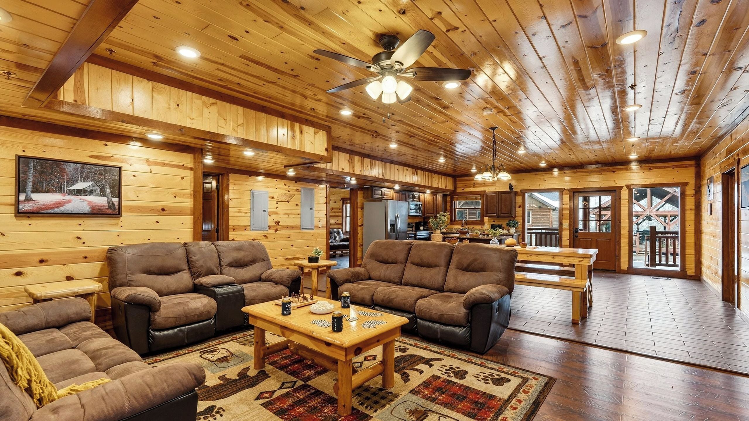 A log cabin interior with a living room, kitchen, and dining area, featuring wooden walls and floors, a ceiling fan, and a large window.