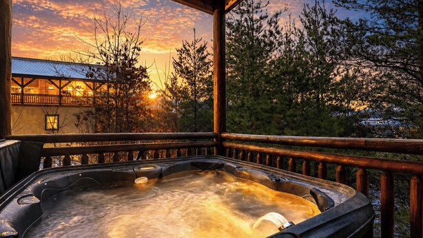 A hot tub with a wooden deck and railing, surrounded by trees and a building with a sloped roof. - Green Mountain Lodge by Avantstay Group Retreat Indoor Pool + Hot Tub + Game Room Pigeon Forge + Near Dollywood (Sevierville)