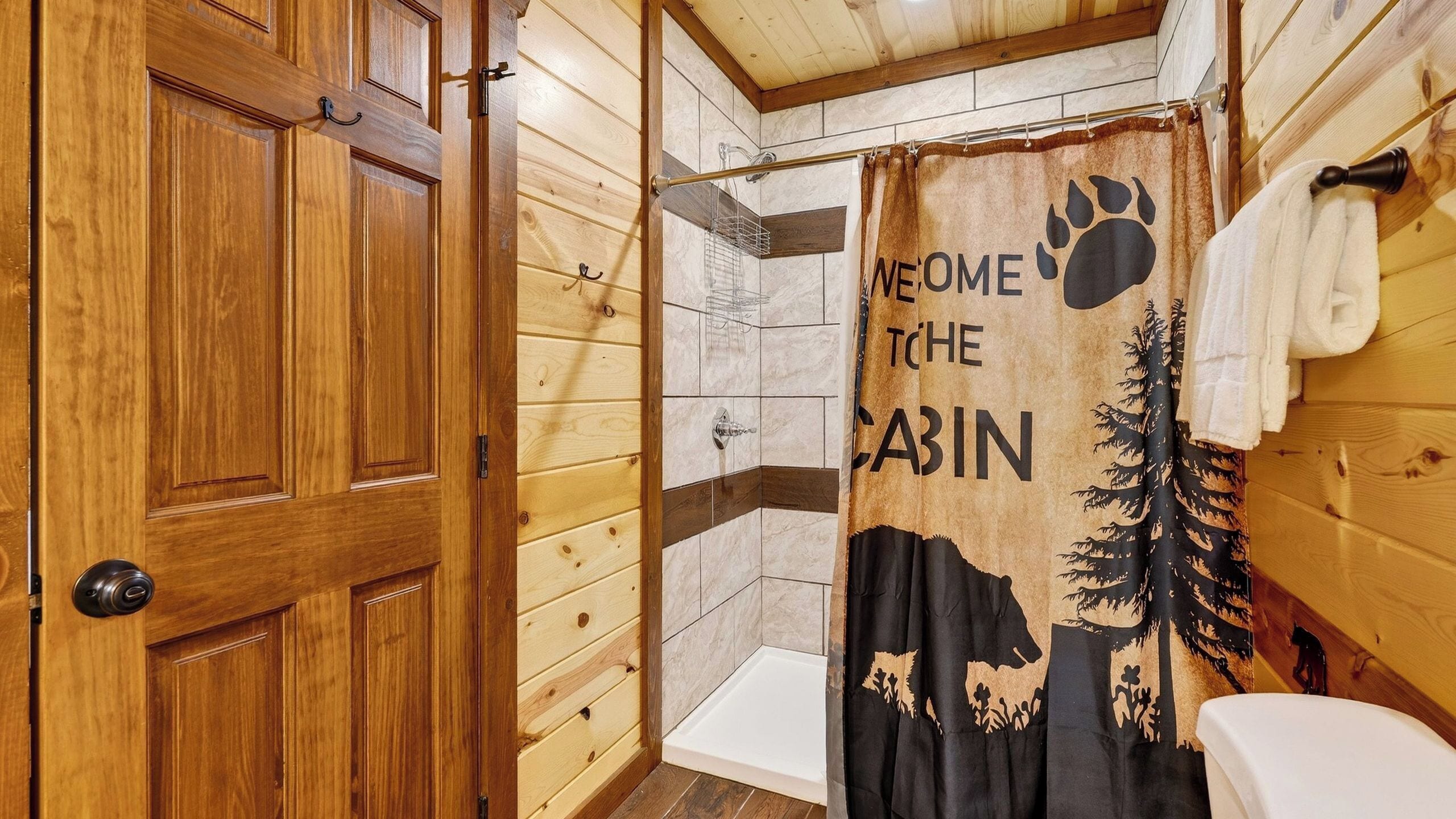A wooden cabin bathroom with a shower, a wooden door, and a 'Welcome to the Cabin' shower curtain.