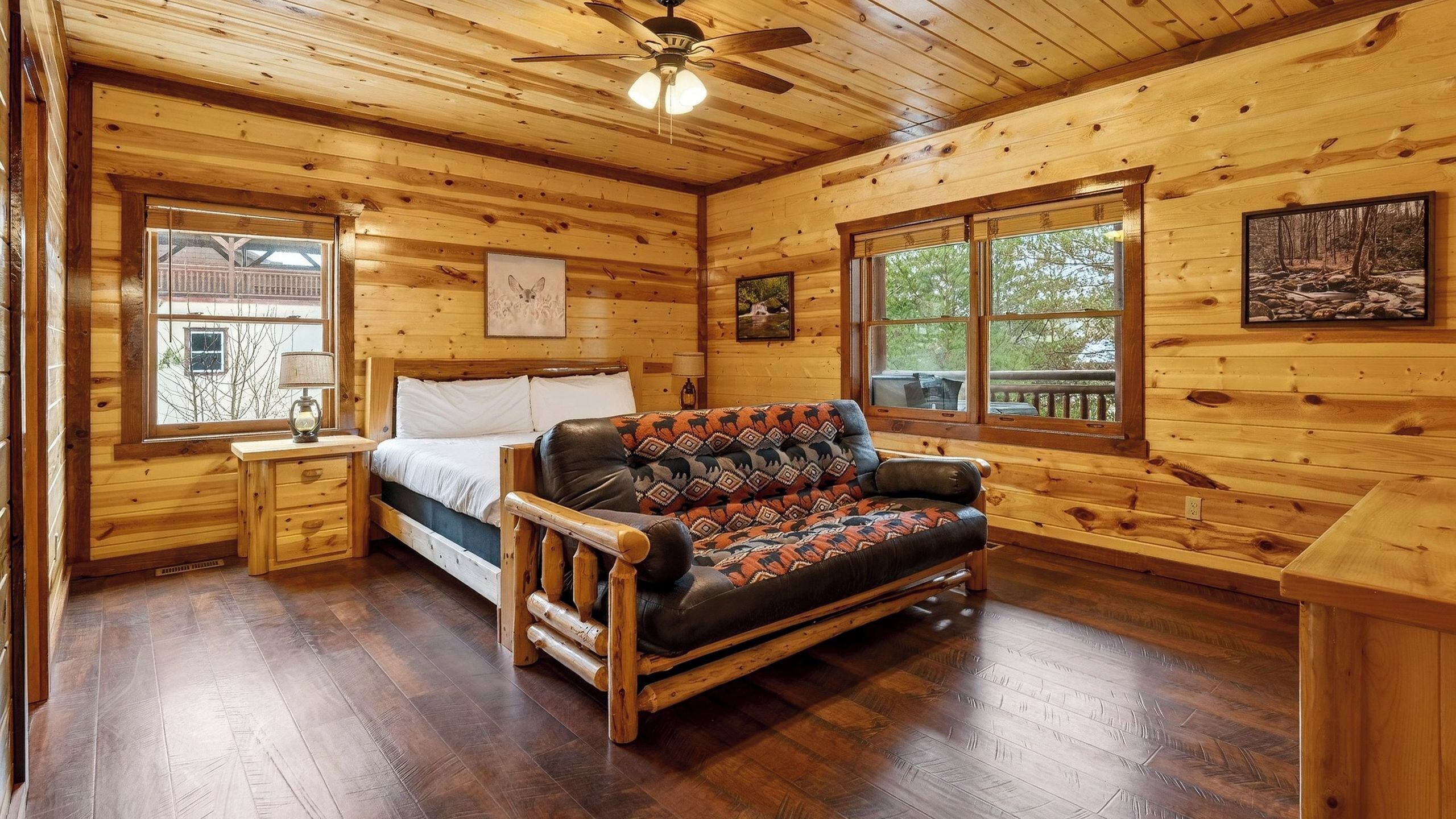 Interior de una cabaña de madera con una cama, un sofá y ventanas amplias.
