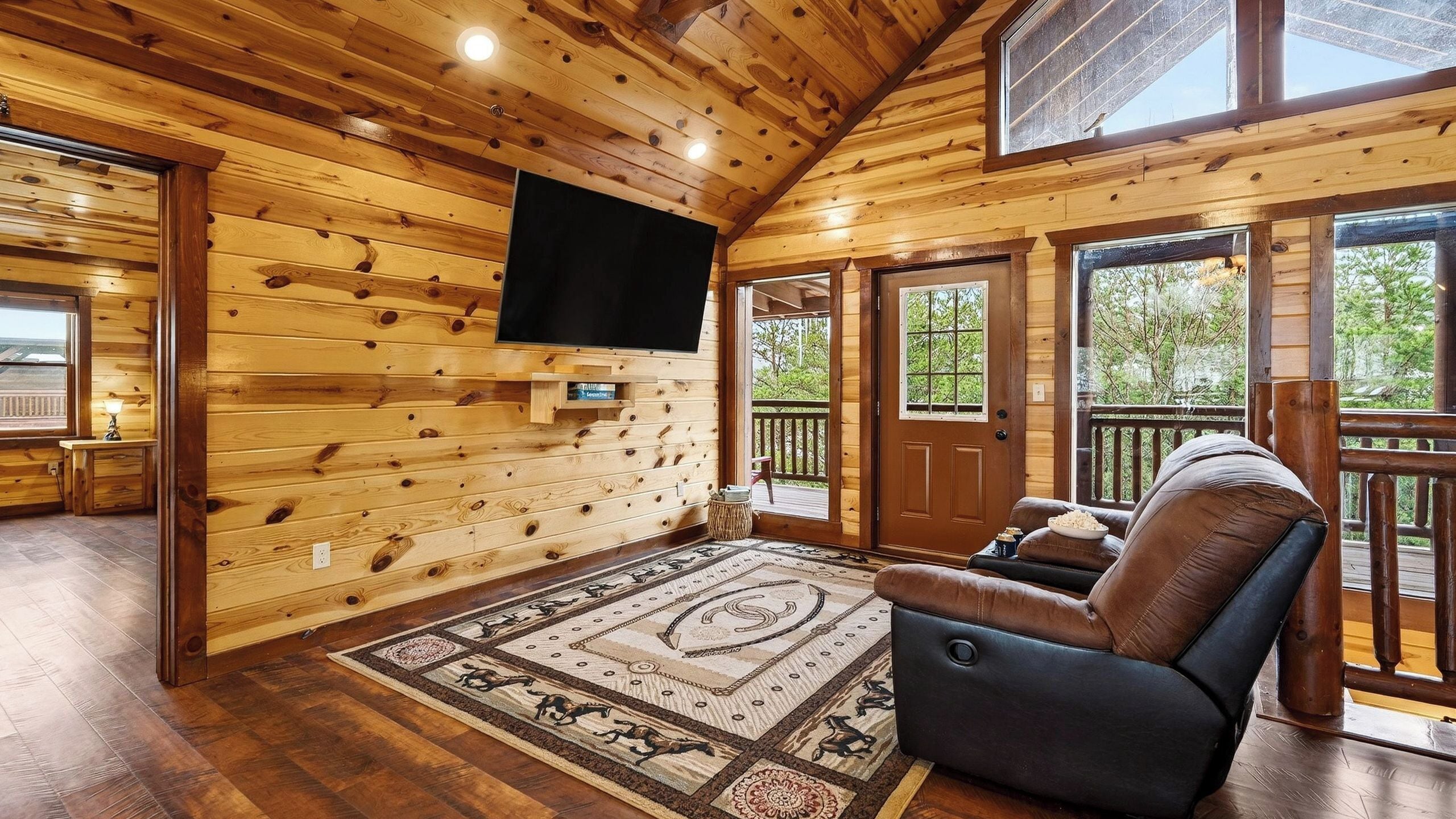 Een houten cabin-interieur met een televisie aan de muur, een groot vloerkleed met een centraal patroon en een leren fauteuil.