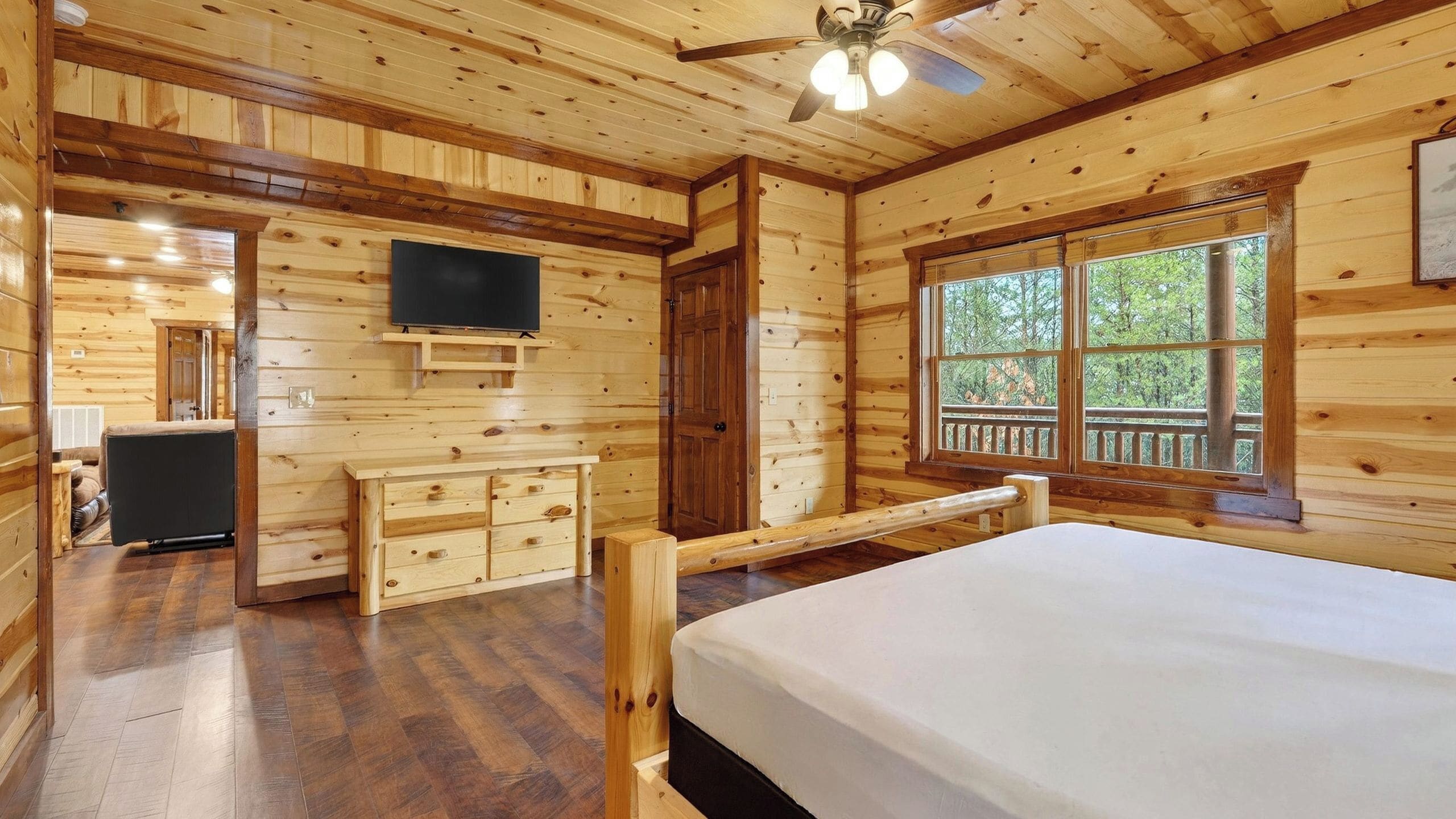 A wooden cabin room with a bed, a TV, a dresser, and a window with a view of trees.