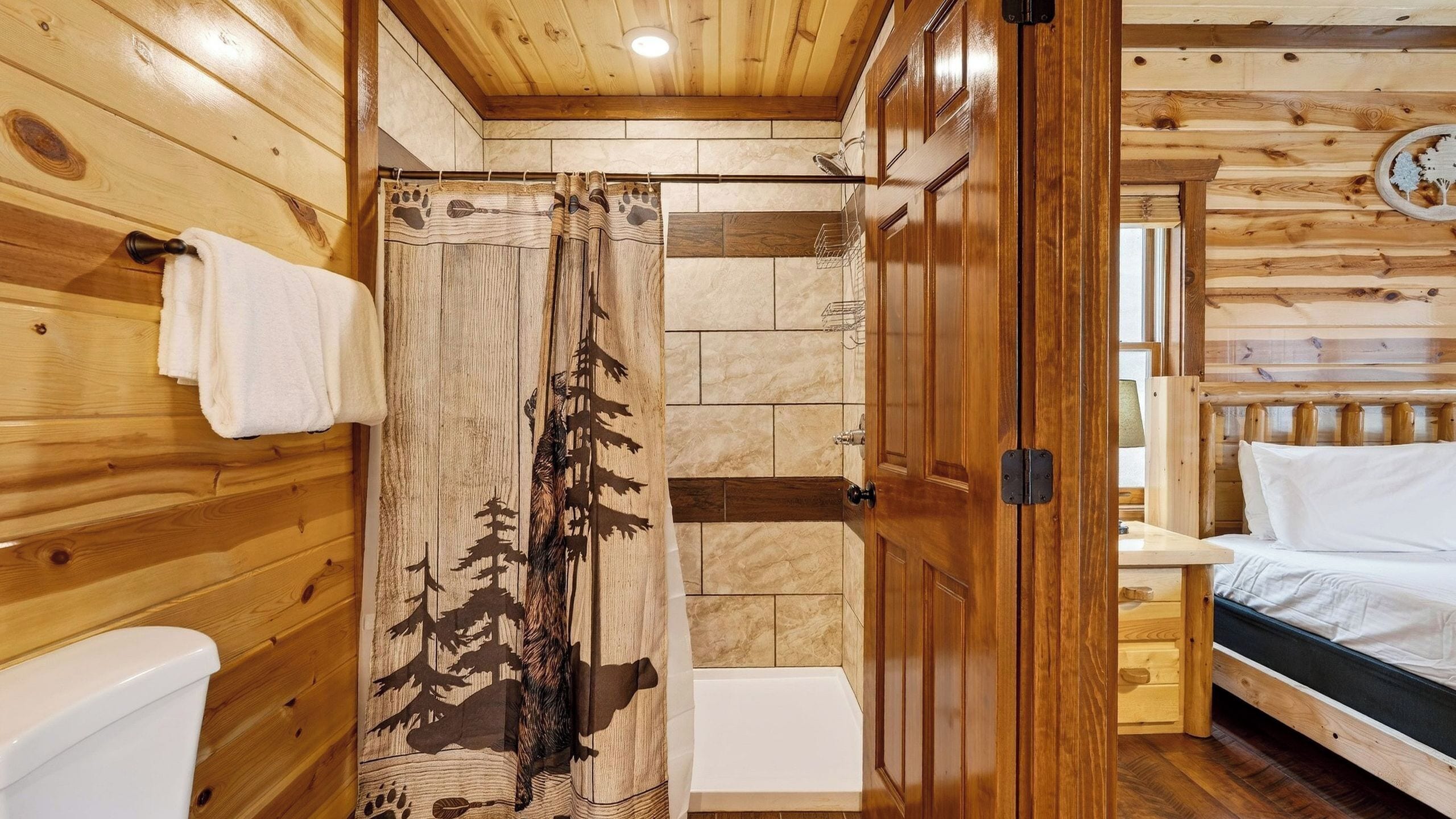 L’intérieur d’un chalet en bois est doté d’une douche avec un rideau orné d’un motif de forêt, d’un lit avec une literie blanche et d’une tête de lit en bois.