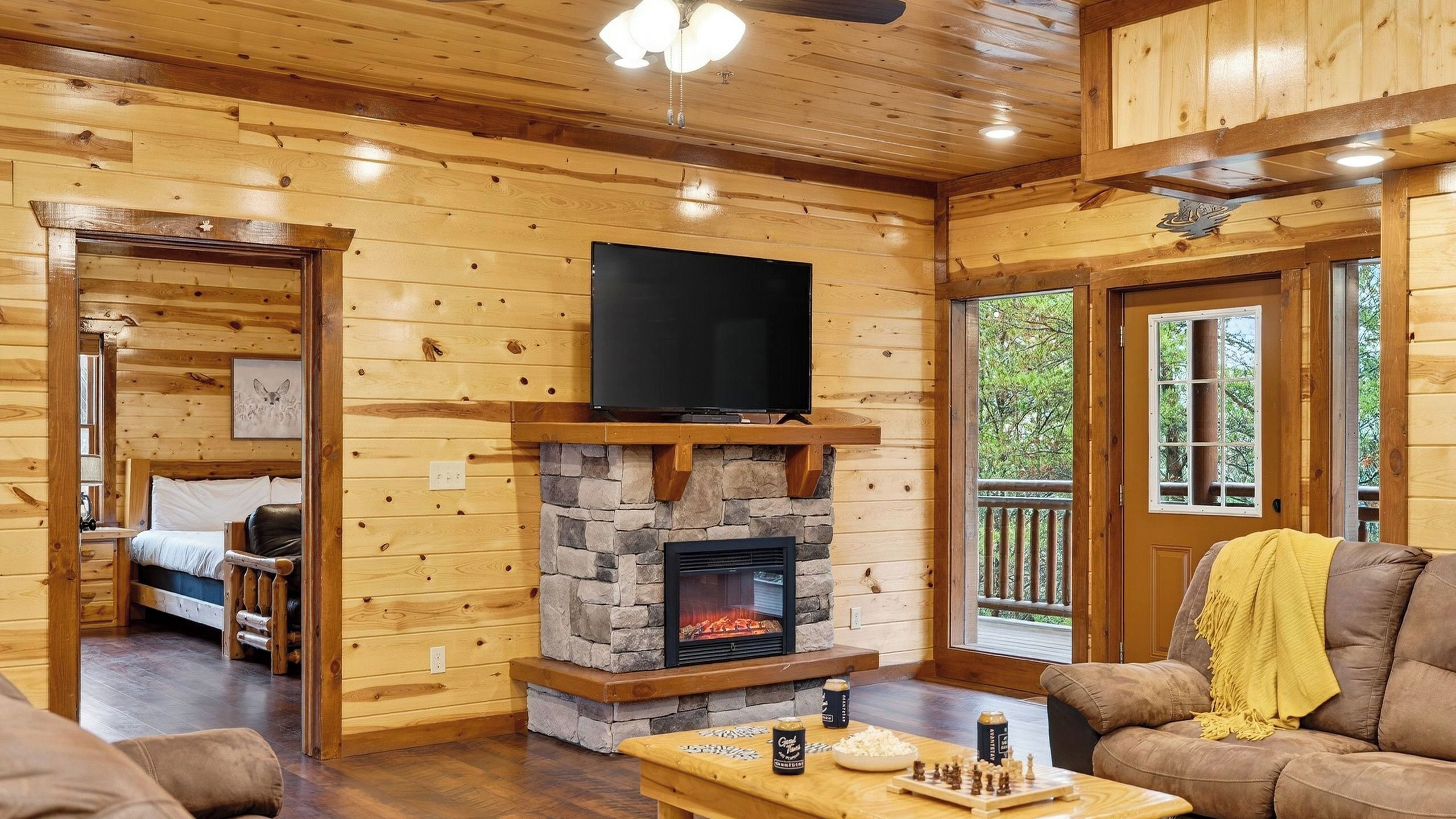 A log cabin interior with a stone fireplace, a television mounted above it, and a cozy seating area with a yellow throw blanket.