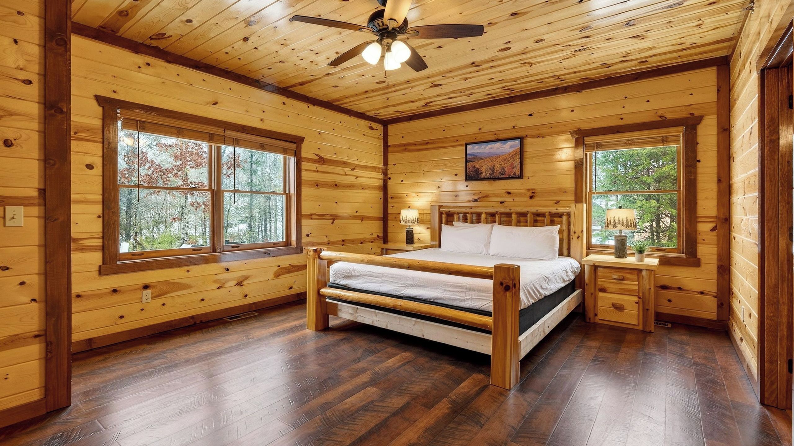 Une chambre à coucher située dans une cabane en bois, comprenant un lit, deux fenêtres, un ventilateur de plafond et une table de chevet avec une lampe.