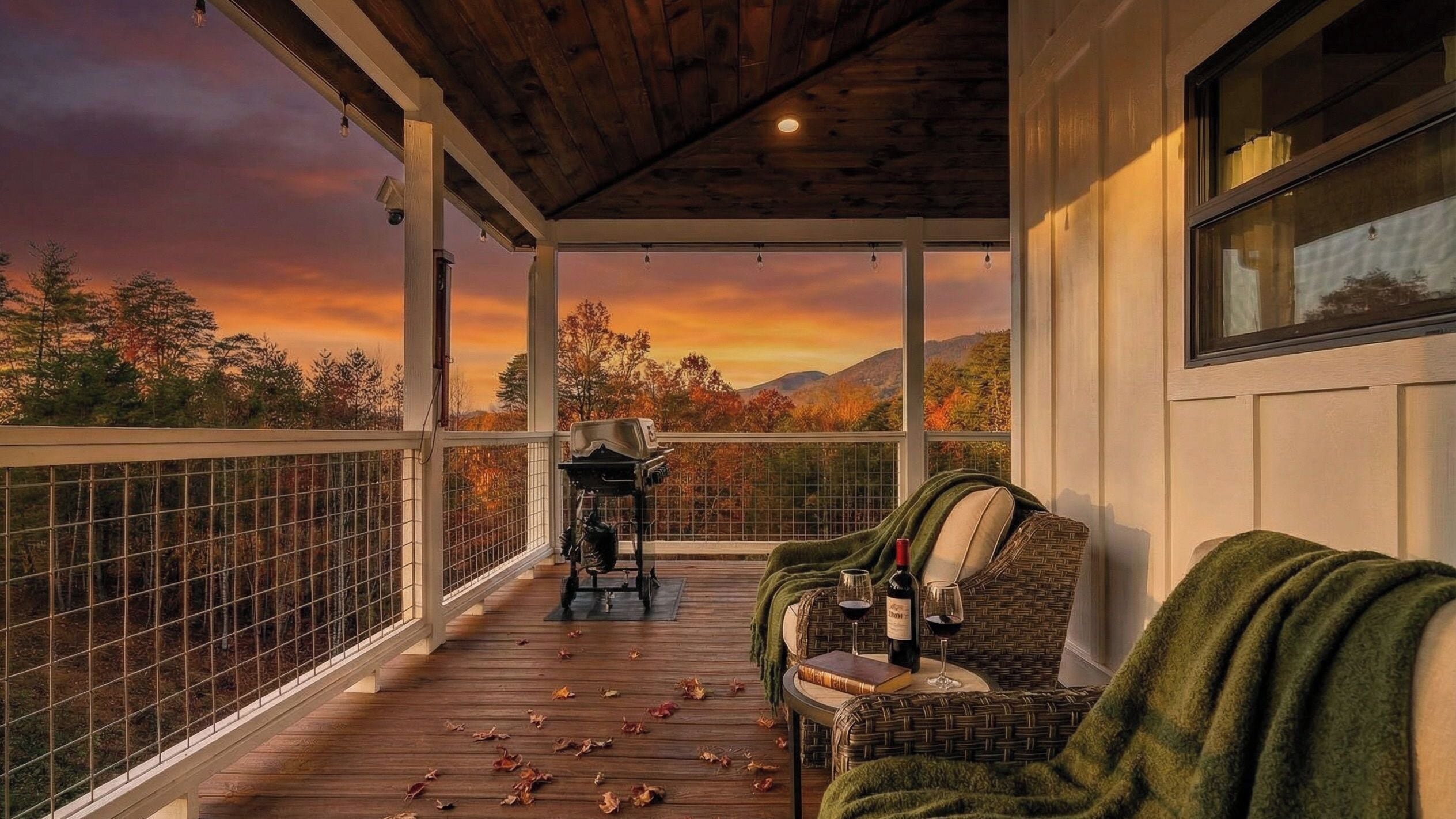 A covered porch with a view of autumn trees and a sunset. There is a grill, a wicker chair, and a table with a book and wine glasses.