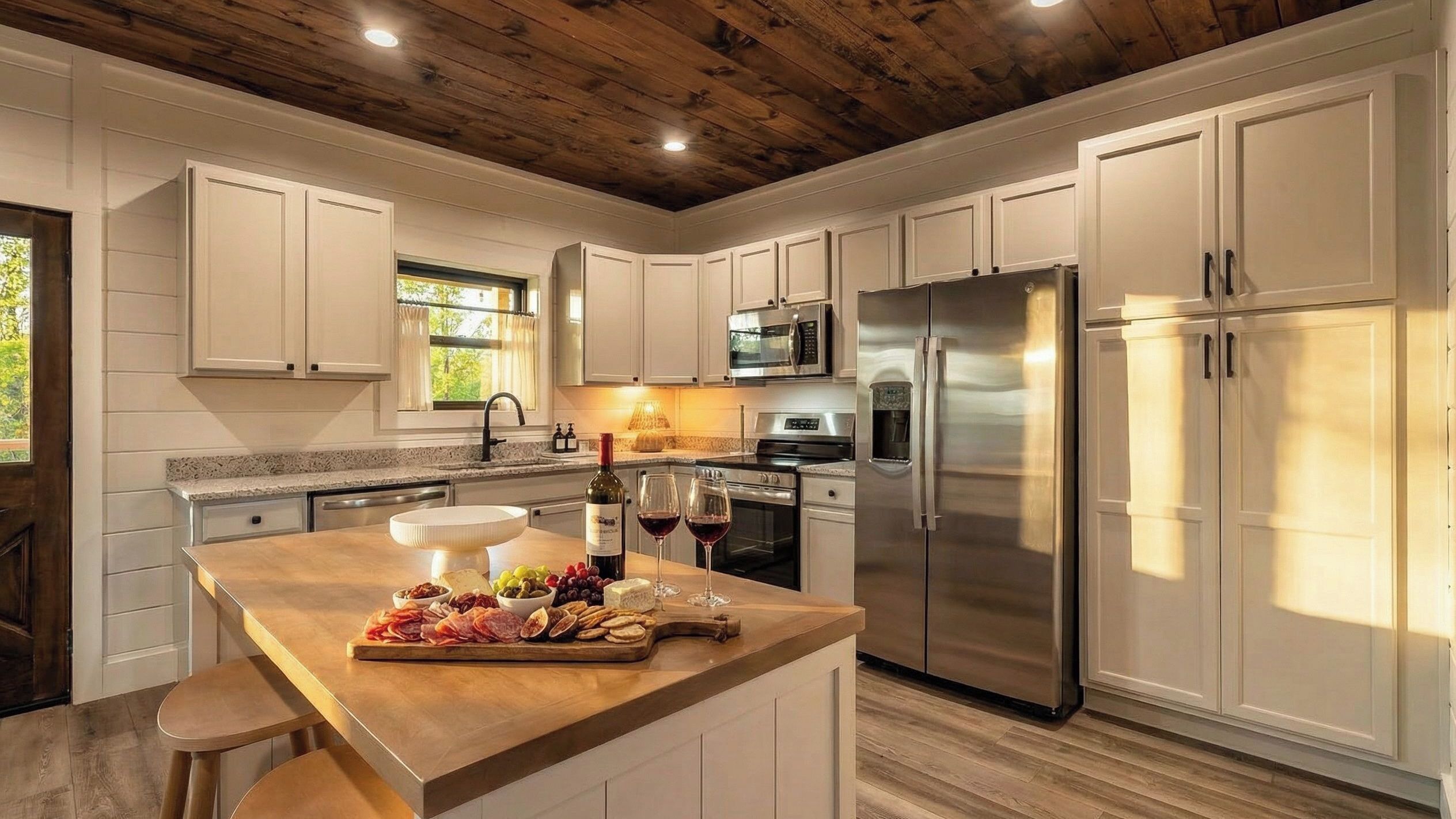 A modern kitchen with white cabinetry, stainless steel appliances, and a wooden island with food and wine.