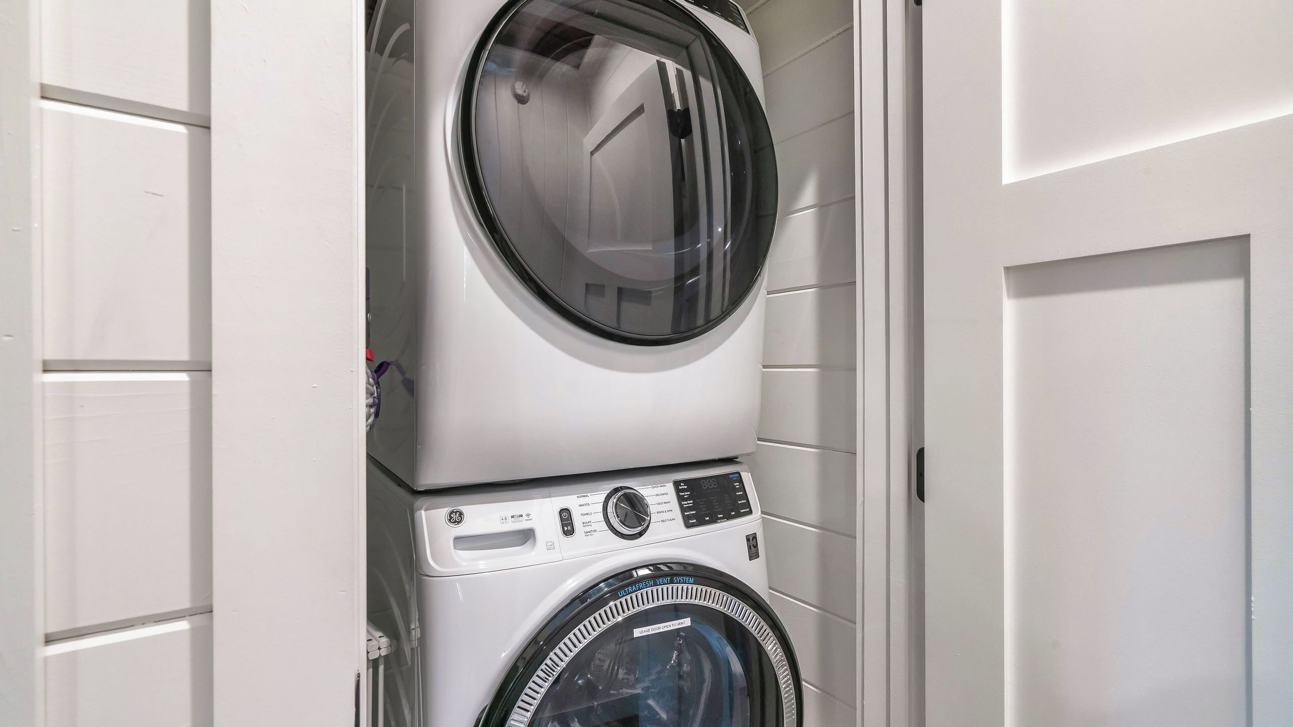 Une machine à laver et un sèche-linge superposés dans une buanderie, avec un mur recouvert de carreaux blancs et une porte.