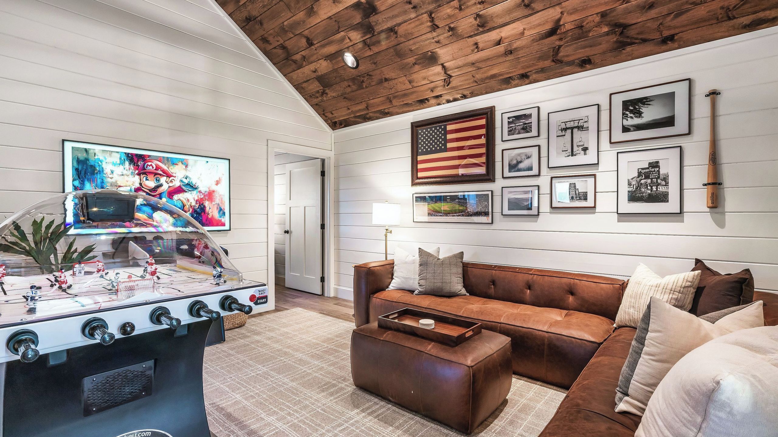 A cozy living room with a foosball table, a brown leather couch, and a wall adorned with framed pictures and an American flag.