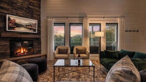 A cozy living room with a fireplace, two armchairs, a green sofa, and large windows with a view of trees and mountains. - White Pines by Avantstay Family Retreat Game Room + Wraparound Porch + Hot Tub (Sevierville)