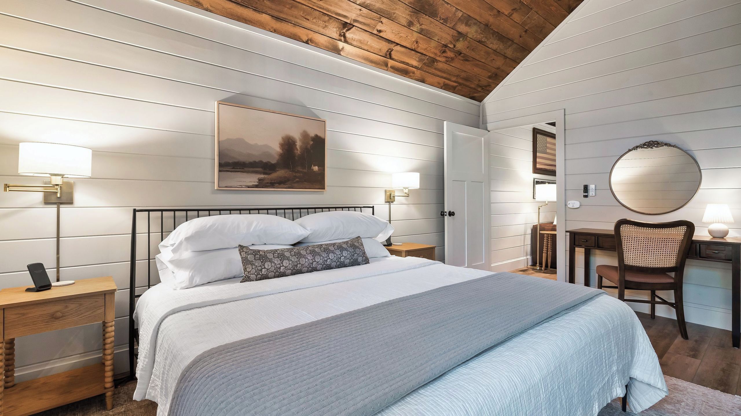 A bedroom with a bed, bedside tables, a desk, a chair, a mirror, and a painting on the wall.