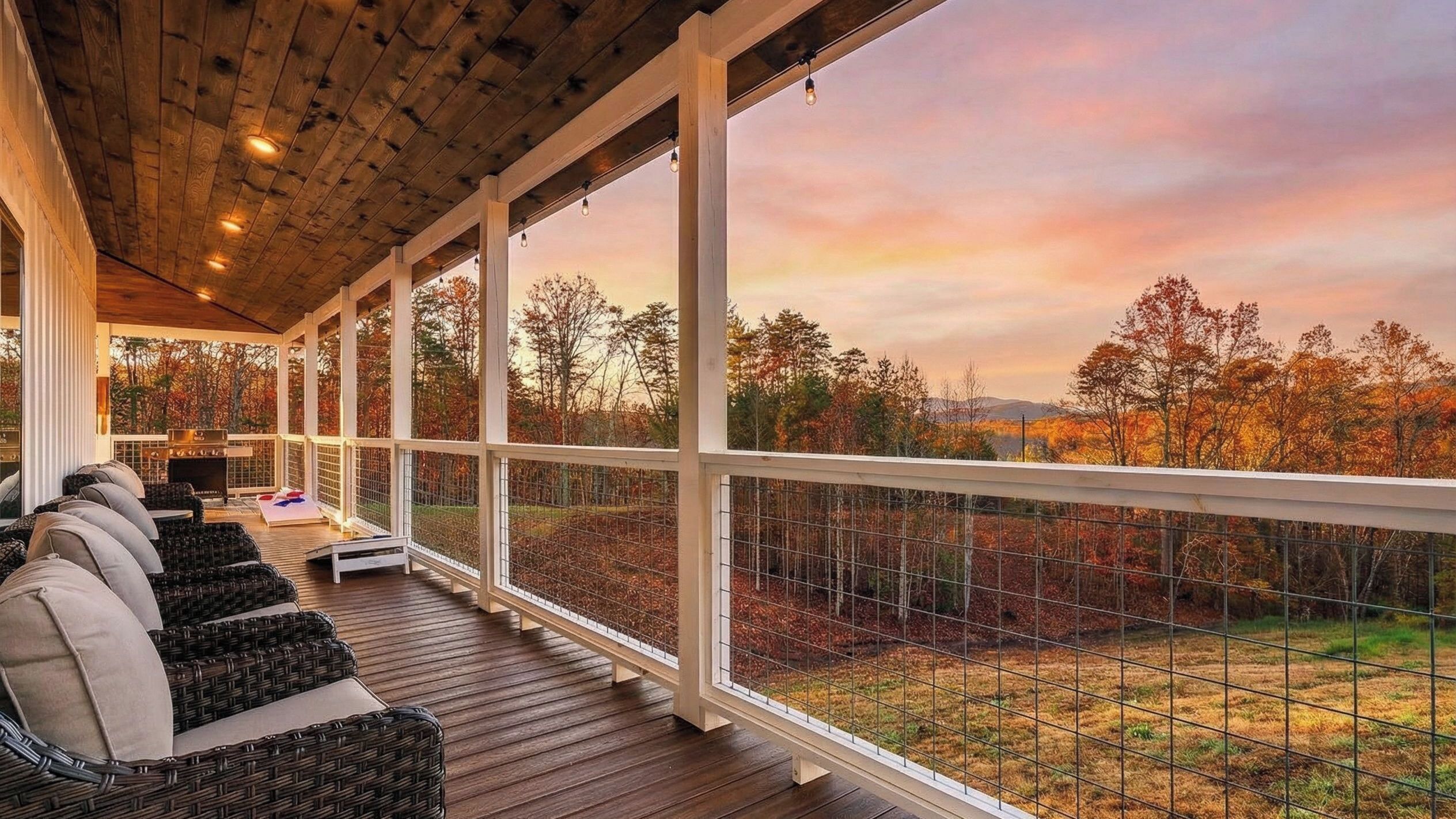 A covered outdoor seating area with wicker furniture and a grill, overlooking a forested landscape.