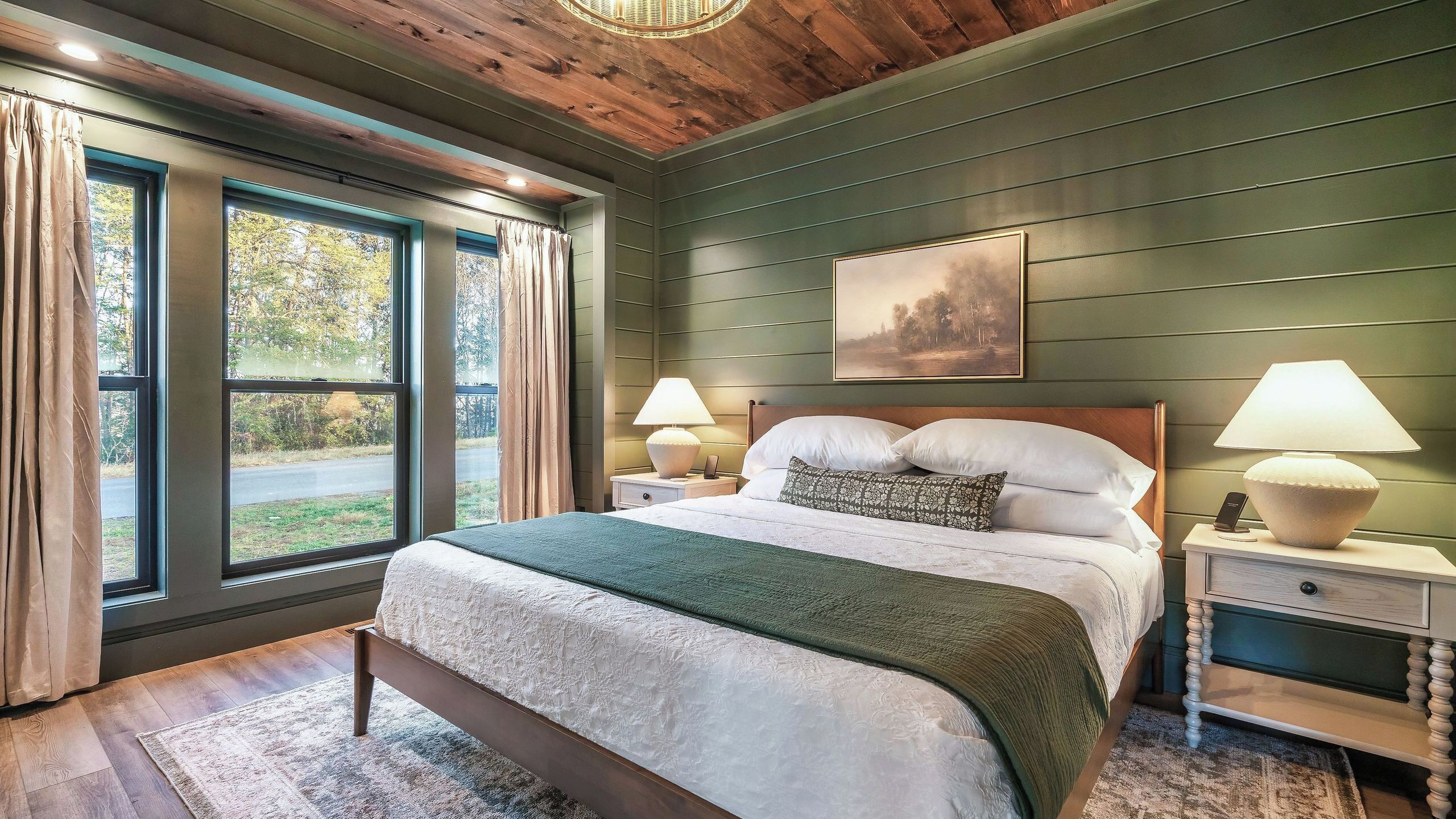 Quarto com uma cama grande, duas luminárias de cabeceira, uma mesa de cabeceira, um quadro na parede e vista para o exterior pelas janelas.