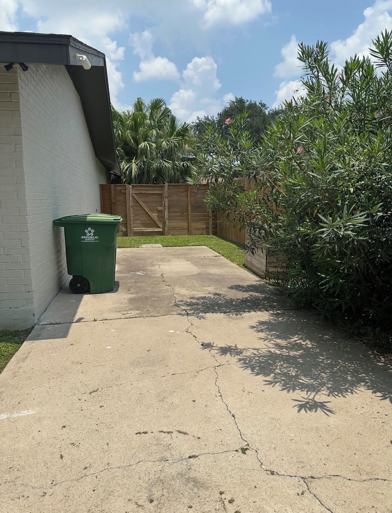 A concrete pathway leading to a wooden fence, a green trash bin, and a white building with a black roof.