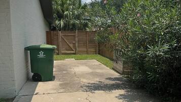 A concrete pathway leading to a wooden fence, a green trash bin, and a white building with a black roof.