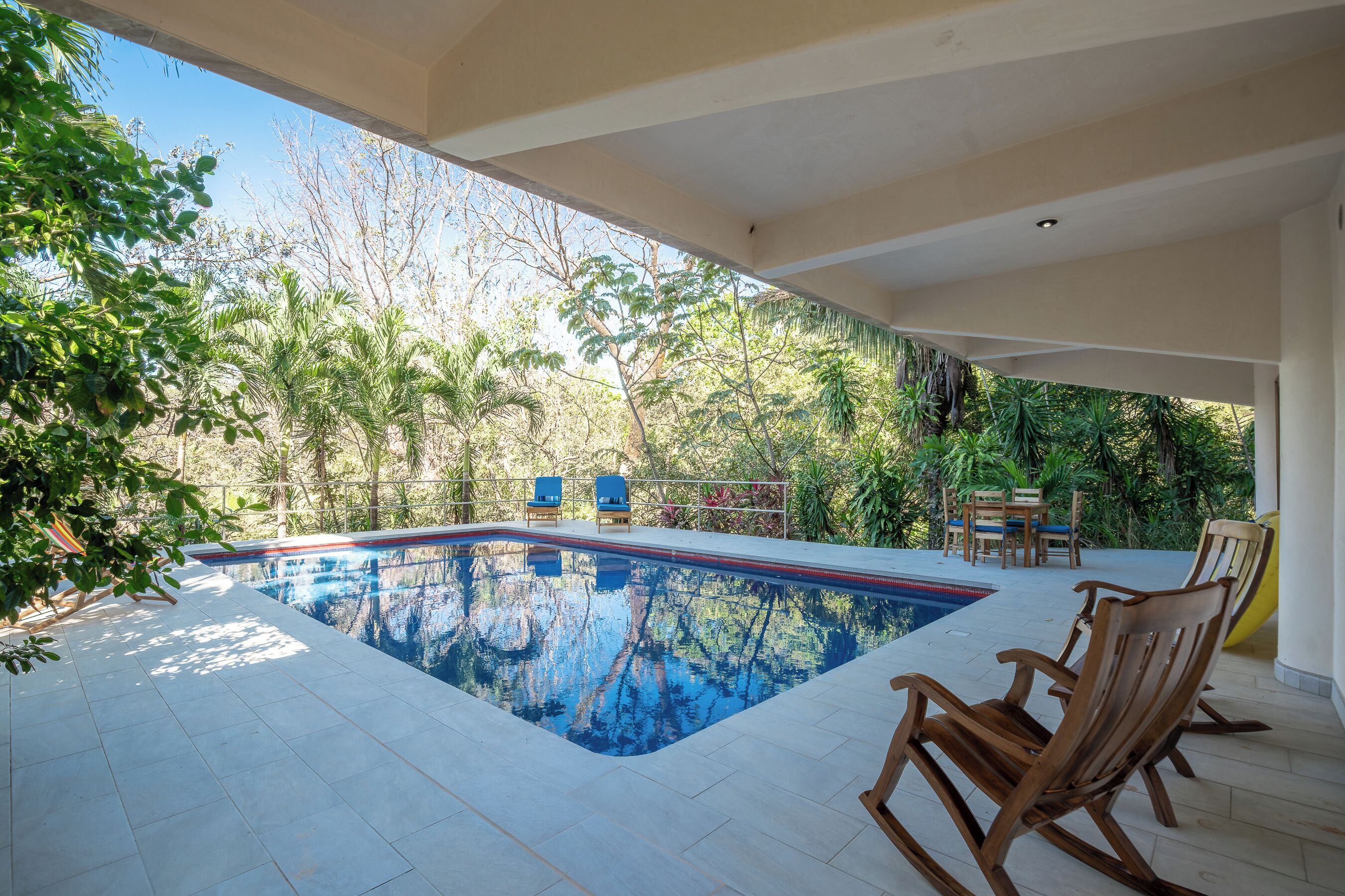 A swimming pool with tiled flooring, surrounded by wooden chairs and tables, with a view of trees and greenery.
