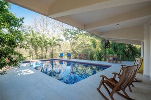 A swimming pool with tiled flooring, surrounded by wooden chairs and tables, with a view of trees and greenery. - Stunning Ocean View at Kahakai House in Guiones (Nosara)