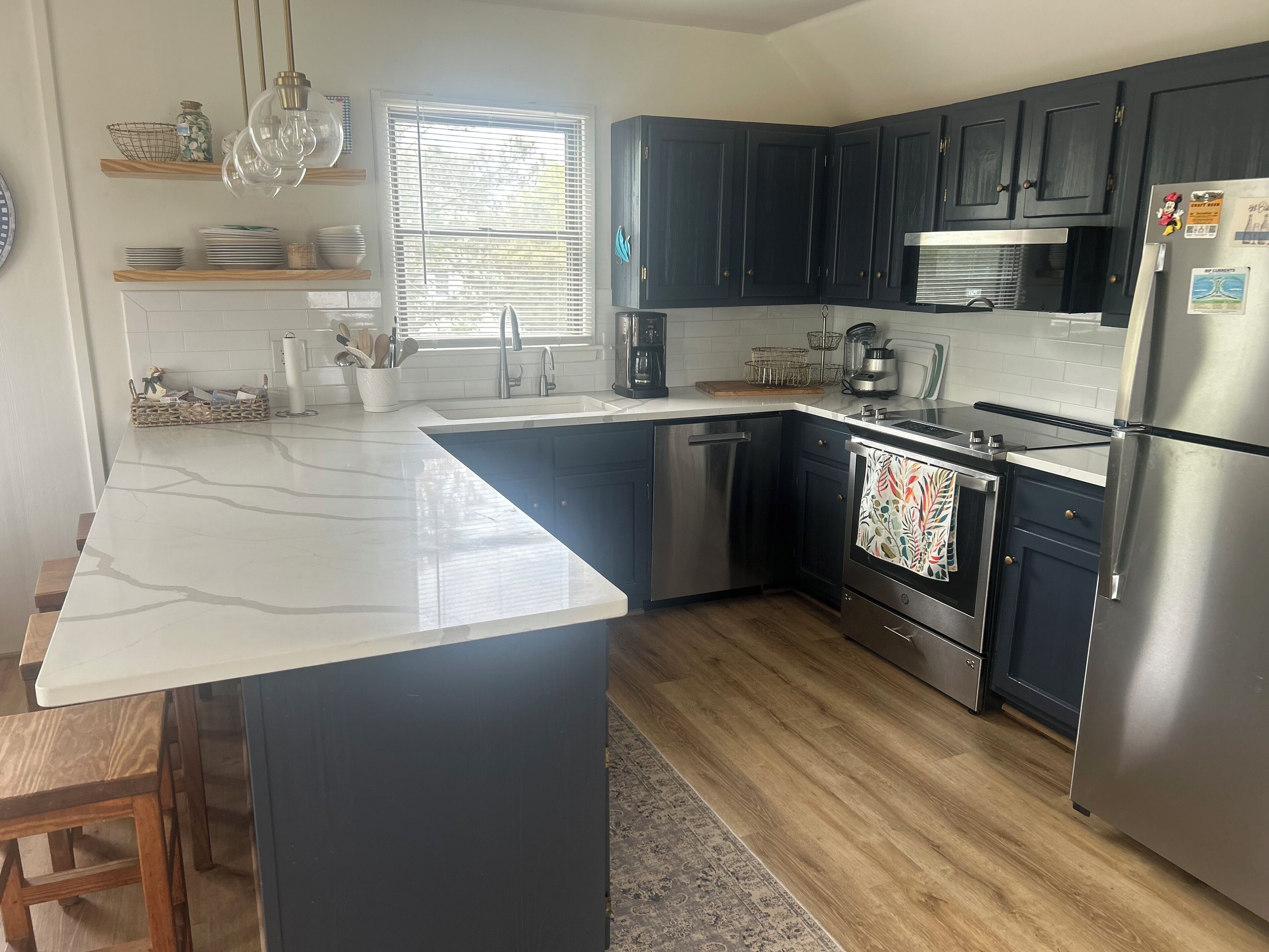 A modern kitchen with dark cabinets, a white marble island, stainless steel appliances, and a window with blinds.